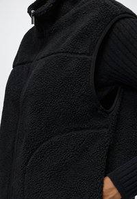 Veste polaire noire texturée avec fermeture éclair à l’avant portée sur un haut noir côtelé à manches longues, main partiellement visible dans la poche.