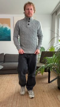 Grijze kabelgebreide trui met een hoge kraag en drie donkere knopen. Draagt zwarte broek en lichte sneakers. Op de achtergrond zijn planten zichtbaar.