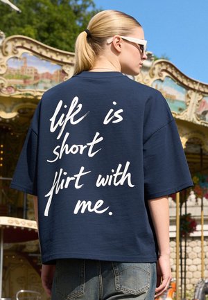Giovane donna che indossa una camicia blu navy con testo bianco "life is short flirt with me," in piedi vicino a una giostra decorativa in una giornata di sole.