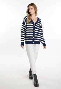 Cardigan a righe blu navy e crema, con scollo a V, maniche lunghe, bottoni frontali e polsini a coste. Indossato sopra pantaloni chiari e stivali neri.