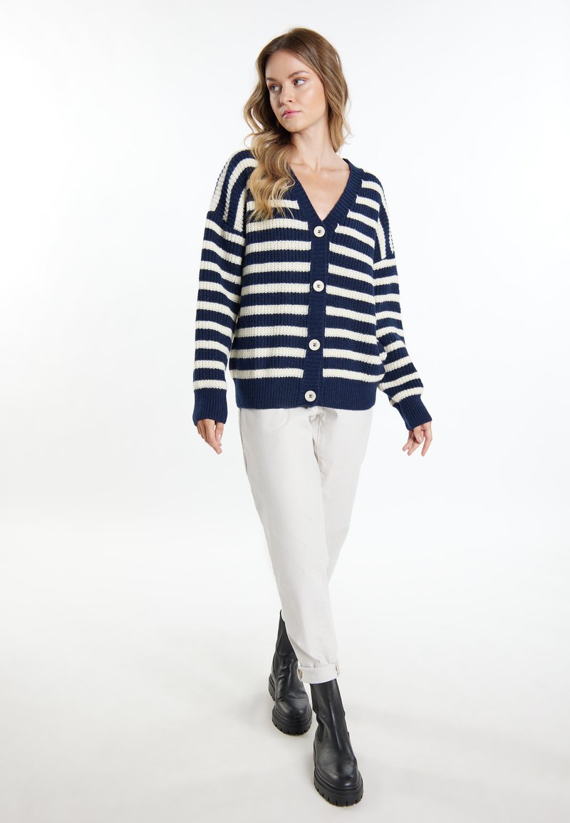 Cardigan a righe blu navy e crema, con scollo a V, maniche lunghe, bottoni frontali e polsini a coste. Indossato sopra pantaloni chiari e stivali neri.