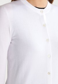 Bílý cardigan vyrobený z lehké pleteniny s kulatým výstřihem, se zapínáním na knoflíky a jemnou strukturou.