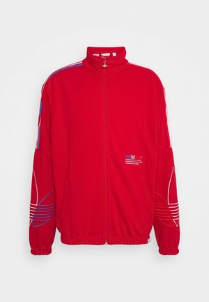 Czerwona kurtka sportowa na pełny zamek z wysokim kołnierzem, z niebiesko-białymi paskami na ramionach i dużymi konturowanymi logo koniczyny na rękawach.