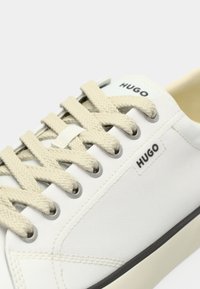 Bela platnena športna obutev z bež vezalkami in črnim yazkom "HUGO" blizu vezalk in na jezičku.