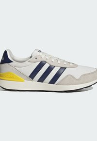 Baskets Adidas blanches et grises avec trois bandes bleu marine et détail jaune au talon, design bas, et lacets blancs.