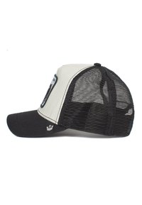 Gorra trucker negra y crema con malla trasera negra, visera curva y un parche en la parte frontal; presenta un logo en el lateral y detalles de costura.