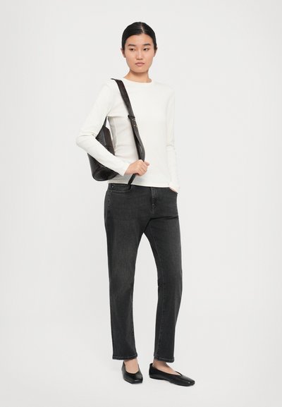 Kvinna som bär en vit långärmad topp, mörkgrå jeans, svarta ballerina-skor och bär en svart läderaxelväska med ett strukturerat mönster.