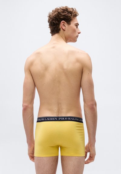 Άνδρας χωρίς πουκάμισο που κοιτάζει μακριά, φοράει κίτρινα μπόξερ με μαύρη μέση στην οποία αναγράφεται «Polo Ralph Lauren», σε απλό λευκό φόντο.