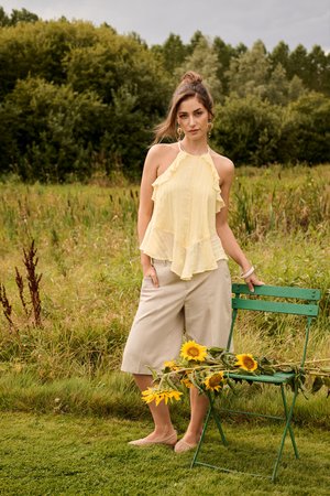 Jonge vrouw in gele top met ruches en beige korte broek staat bij een groene stoel met zonnebloemen in een grasveld onder een bewolkte hemel.