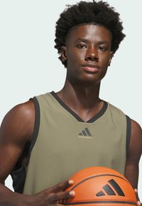 Oliivinvihreä koripallopaita mustilla yksityiskohdilla ja kultaisella logolla. Pelaaja pitää oranssia koripalloa. Sujuva kangas, urheilullinen muoto.