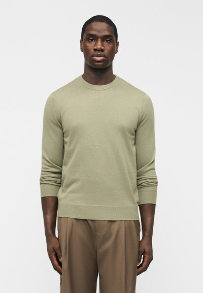 Homme debout, face à l'avant, portant un pull col rond vert clair et un pantalon marron, sur un fond blanc uni.