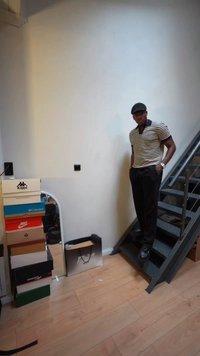 Άνδρας με ριγέ πουκάμισο και μαύρο παντελόνι στέκεται σε μεταλλική σκάλα, δίπλα σε στοιβαγμένα κουτιά παπουτσιών και έναν δαπέδου καθρέφτη σε δωμάτιο με ξύλινο πάτωμα.