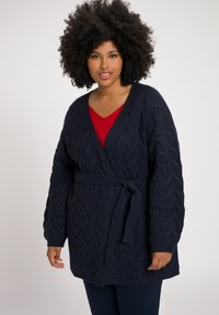 Donna con capelli ricci che indossa un top rosso e un cardigan a girocollo blu navy con cintura, in piedi contro uno sfondo bianco uniforme.