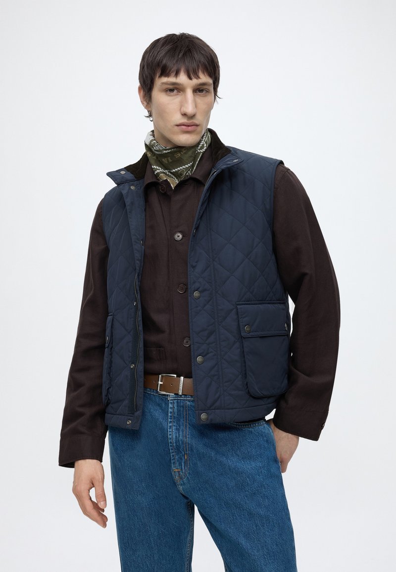 Mand iført en marineblå vatteret vest over en mørkebrun skjorte, blå jeans, brun bælte og et grønt mønstret halstørklæde, står mod en neutral baggrund.