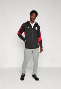 Svart zip-up hettegenser med røde detaljer og gull logoer, sammen med grå joggebukser. Outfitten har et moderne atletisk design og en komfortabel passform.