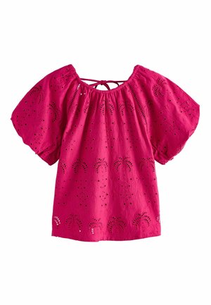Femeie cu păr creț închis la culoare, purtând o bluză roz cu perforații și pantaloni scurți gri brodați cu flori, stă pe o plajă la apus.