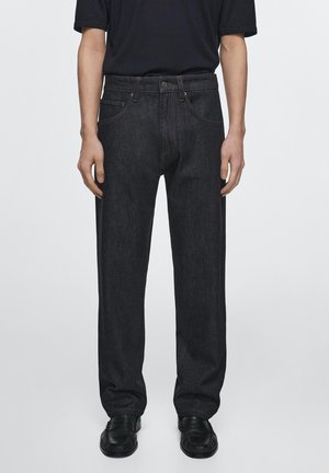 Person trägt schwarze Denim-Jeans mit geradem Bein, schwarze Lederschuhe und ein schwarzes kurzärmeliges Hemd und steht vor einem schlichten Hintergrund.