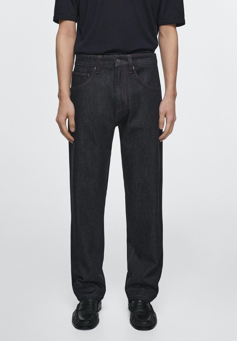 Person trägt schwarze Denim-Jeans mit geradem Bein, schwarze Lederschuhe und ein schwarzes kurzärmeliges Hemd und steht vor einem schlichten Hintergrund.