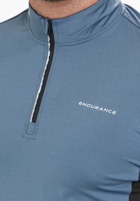 Helles blaues Sportshirt mit einem halben Reißverschluss am Hals, schwarzen Seiteneinsätzen und einem weißen "ENDURANCE"-Logo auf der Vorderseite, aus glattem, atmungsaktivem Material gefertigt.