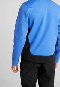 Blauwe en zwarte jas met een gladde textuur, voorzien van lange mouwen en een gebogen ontwerp aan de achterkant. Inclusief een ritsvak.