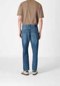 Vaaleanruskea t-paita, takanäkymä, yhdistetty sinisiin farkkuihin. Farkuissa on klassinen istuvuus, viisi taskua ja lievää haalistumista. Jalkineet: vaaleanbeige lenkkarit.