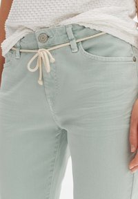 Pantaloni in denim verde chiaro con vita regolabile. Presentano una chiusura con bottone metallico e due tasche frontali, evidenziando una texture liscia.