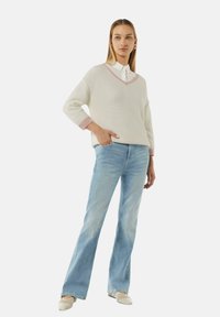 Creme V-hals gebreide trui met roze accenten, gecombineerd met lichtblauwe flared jeans en beige instappers. Lichte textuur en ontspannen pasvorm.