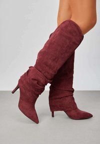 Bottes bordeaux au-dessus du genou en suède doux avec un design ample et une pointe effilée. Présente un talon haut slim pour une élévation supplémentaire.