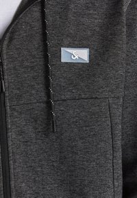 Sudadera con cremallera gris hecha de tejido texturizado, con un bolsillo con cremallera y un pequeño parche de logo azul. Incluye cordones con acentos blancos.