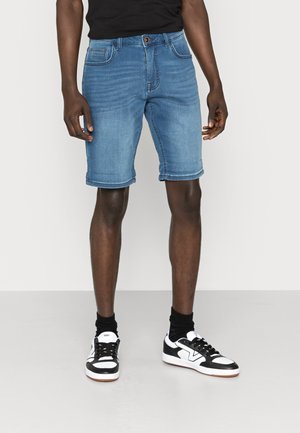 Mand iført blå denimshorts, sort skjorte, sorte sokker og sorte og hvide sneakers, der står mod en ensfarvet baggrund.