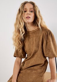 Robe en suède marron avec des manches courtes bouffantes, un coupe ample et un col rond. Texture lisse, détails minimaux et longueur genoux.