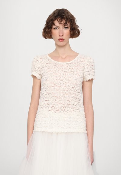 Haut en dentelle blanc cassé avec des manches courtes, texture festonnée et encolure arrondie, superposé sur une jupe en tulle transparent.