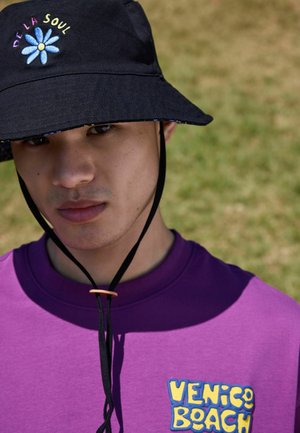 Junger Mann trägt einen schwarzen Eimerhut mit einer blauen Blume und dem Schriftzug "De La Soul" sowie ein lila Shirt mit gelbem "Venice Beach"-Logo.