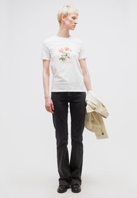 Άτομο με κοντά ξανθά μαλλιά που φοράει λευκό μπλουζάκι με γραφικό σχέδιο λευκού τριαντάφυλλου, μαύρα τζιν, μαύρα παπούτσια, κρατώντας μπεζ μπουφάν, κοιτάζει προς τα πλάγια.