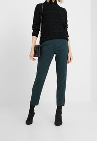 Pull noir côtelé avec des boucles texturées, associé à un pantalon sarouel teal et des bottines noires, portant un petit sac à main noir.