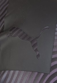 Svarta skor med en upphöjd svart Puma-logotyp på en texturerad bakgrund med lila och svarta ränder. Slätt material med subtil glans.