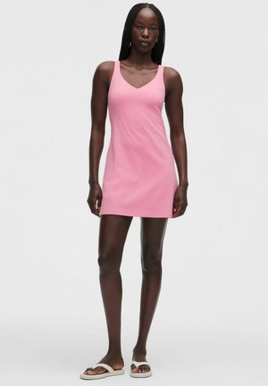 Mujer con largas trenzas que lleva un mini vestido rosa sin mangas y chanclas blancas, de pie frente a un fondo liso de color gris claro.