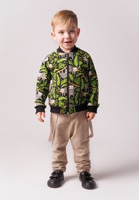 Dziecięca kurtka zapinana na zamek w kolorze czarnym z zielonym wzorem liści i leniwca, zestawiona z beżowymi spodniami i czarnymi trampkami z rzepami.