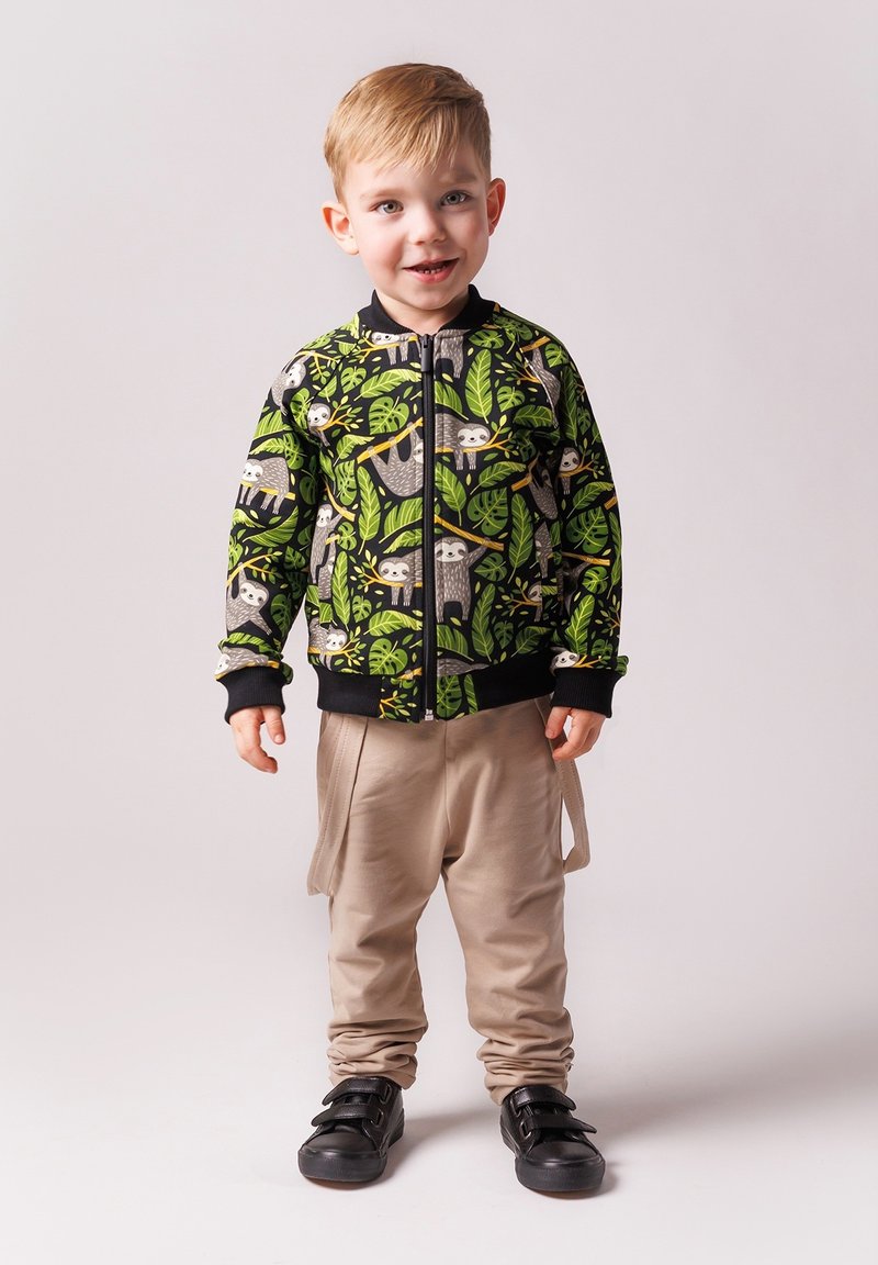 Dziecięca kurtka zapinana na zamek w kolorze czarnym z zielonym wzorem liści i leniwca, zestawiona z beżowymi spodniami i czarnymi trampkami z rzepami.