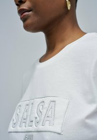 Primer plano de una persona que lleva una camisa blanca con "SALSA" escrito en pedrería y un pendiente de aro dorado visible en una oreja.