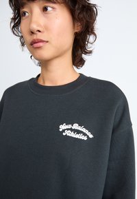 Mørkegrå hettegenser laget av mykt stoff, med en hvit "New Balance Athletics" logo brodert på venstre bryst.
