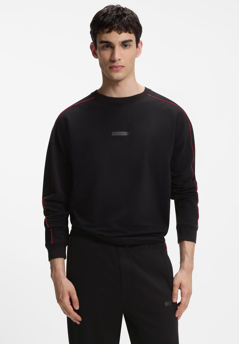 Jeune homme portant une chemise noire à manches longues et un pantalon avec des détails de passepoil rouge sur un fond blanc uni.