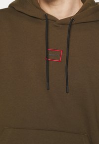 Kapuzensweatshirt aus olivgrünem Stoff mit einem kleinen roten Quadrat-Label, auf dem "HUGO" in Grau steht, schwarzen Kordeln und einer Fronttasche.