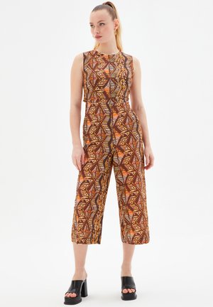 Braun gemusterter Jumpsuit mit geometrischen Designs in Orange, Gelb und Weiß. Ärmelloses Design, verkürzte Hosenbeine, kombiniert mit schwarzen Blockabsatzschuhen.