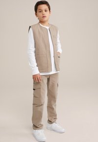 Beige arbetsväst med sidfickor över en vit långärmad skjorta, ihopparad med beiga cargobyxor och vita sneakers. Slät, avslappnad design.