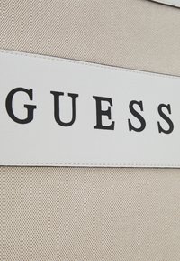 Beżowe tło z tkaniny z wyraźnym białym paneliem skórzanym, na którym widnieje słowo "GUESS" napisane pogrubionymi czarnymi literami. Widoczne czyste szwy.