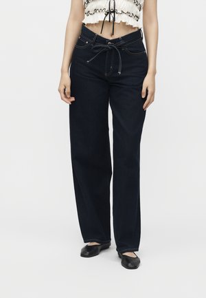 Femme portant un jean bleu foncé à jambes larges avec une ceinture nouée à la taille, un top court blanc et des chaussures plates noires sur un fond uni.