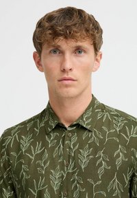 Camicia verde con bottoni, decorata con un motivo a foglie, dotata di colletto, maniche corte e una texture di tessuto morbido. I bottoni sono marroni.