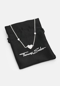 Srebrny naszyjnik z wisiorkiem w kształcie serca o teksturowanej powierzchni i dwoma okrągłymi akcentami, spoczywający na czarnej tkaninowej saszetce z logo "Thomas Sabo".