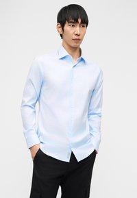 Νέος άνδρας που φοράει γαλάζιο πουκάμισο με κουμπιά, με τα χέρια στις τσέπες μαύρων παντελονιών, κοιτάζοντας ελαφρώς προς τα δεξιά σε απλό λευκό φόντο.